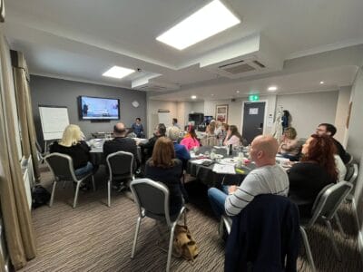Meeting room with people sitting around 3 tables, watching a video as part of a training by One in Four